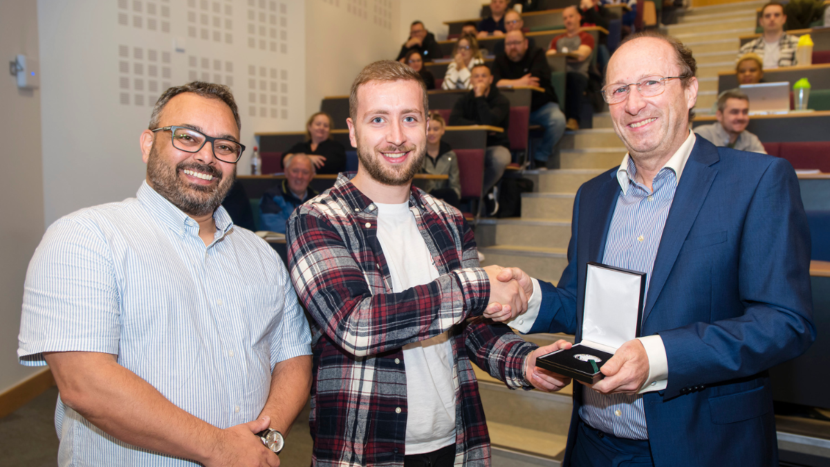 Three men in lecture theatre, one being presented with a medal